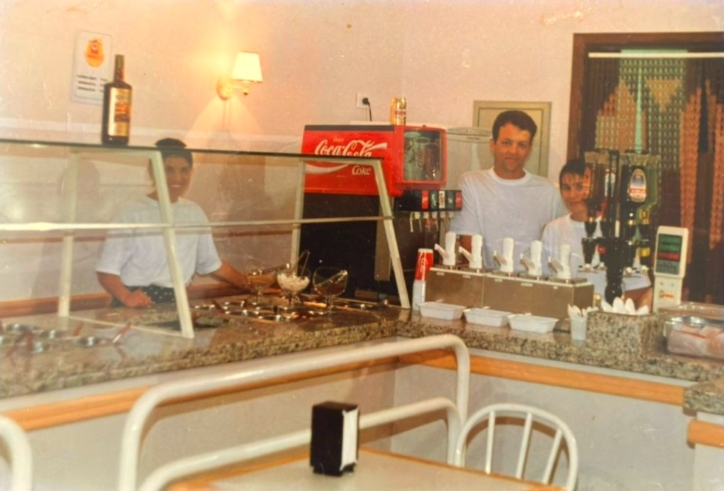 Foto afetiva de três funcionários sorridentes atrás do balcão do Skandallus.